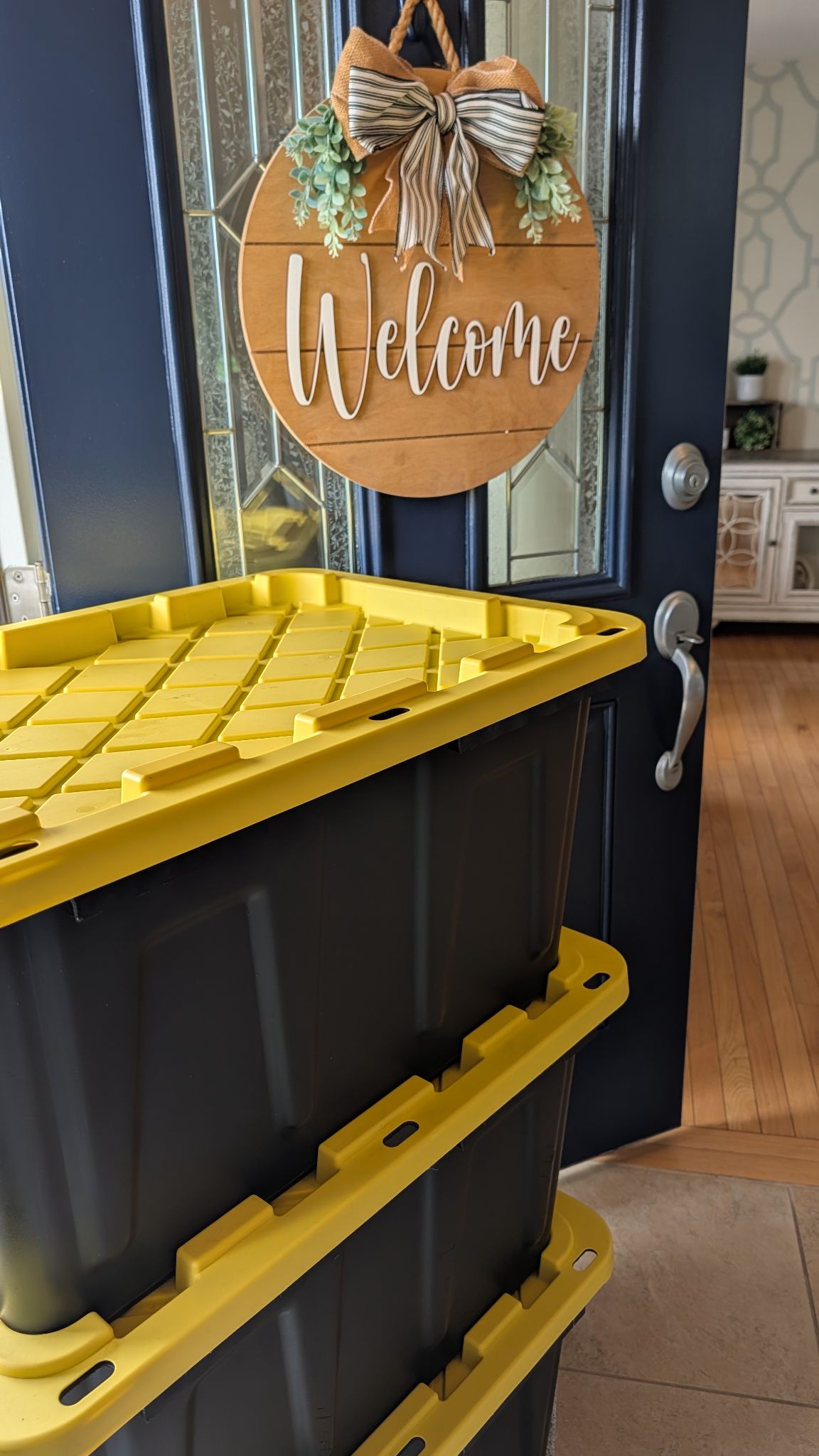 Three heavy-duty black moving totes with yellow lids stacked in an open front doorway beneath a wooden Welcome sign