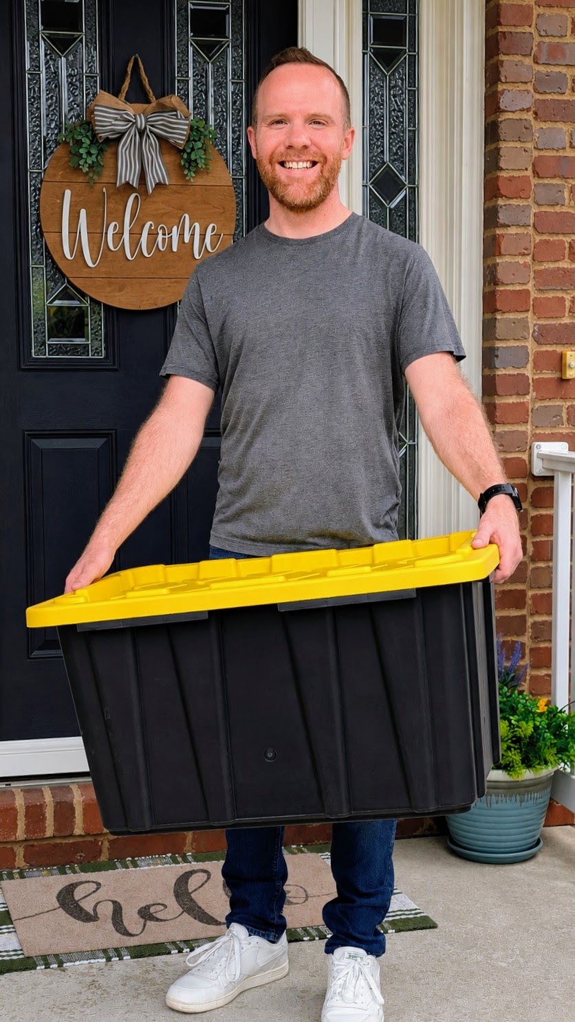 Ryan smiling at a front door, holding a black moving tote with a yellow lid.